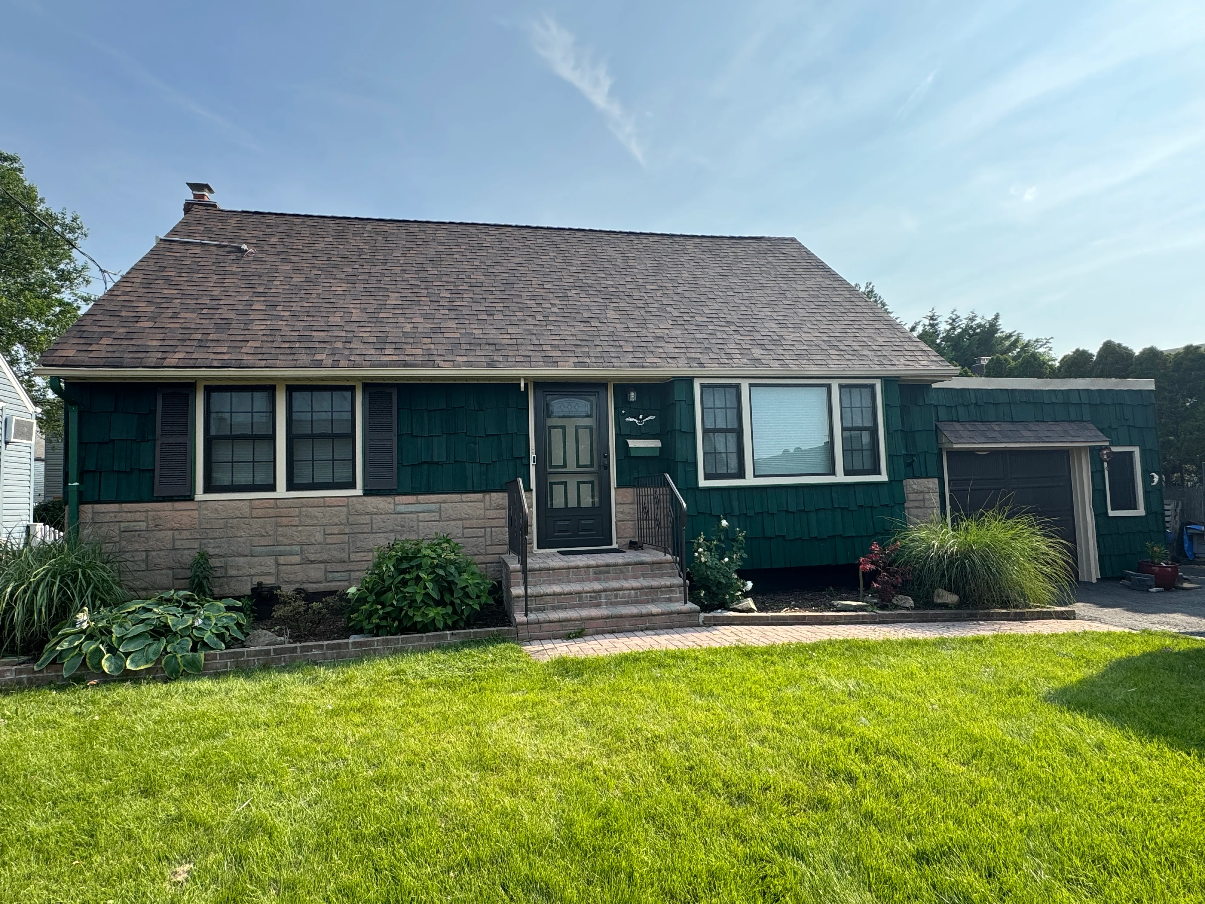 Green shingled cottage with new windows, dark siding, brick base, and manicured front lawn