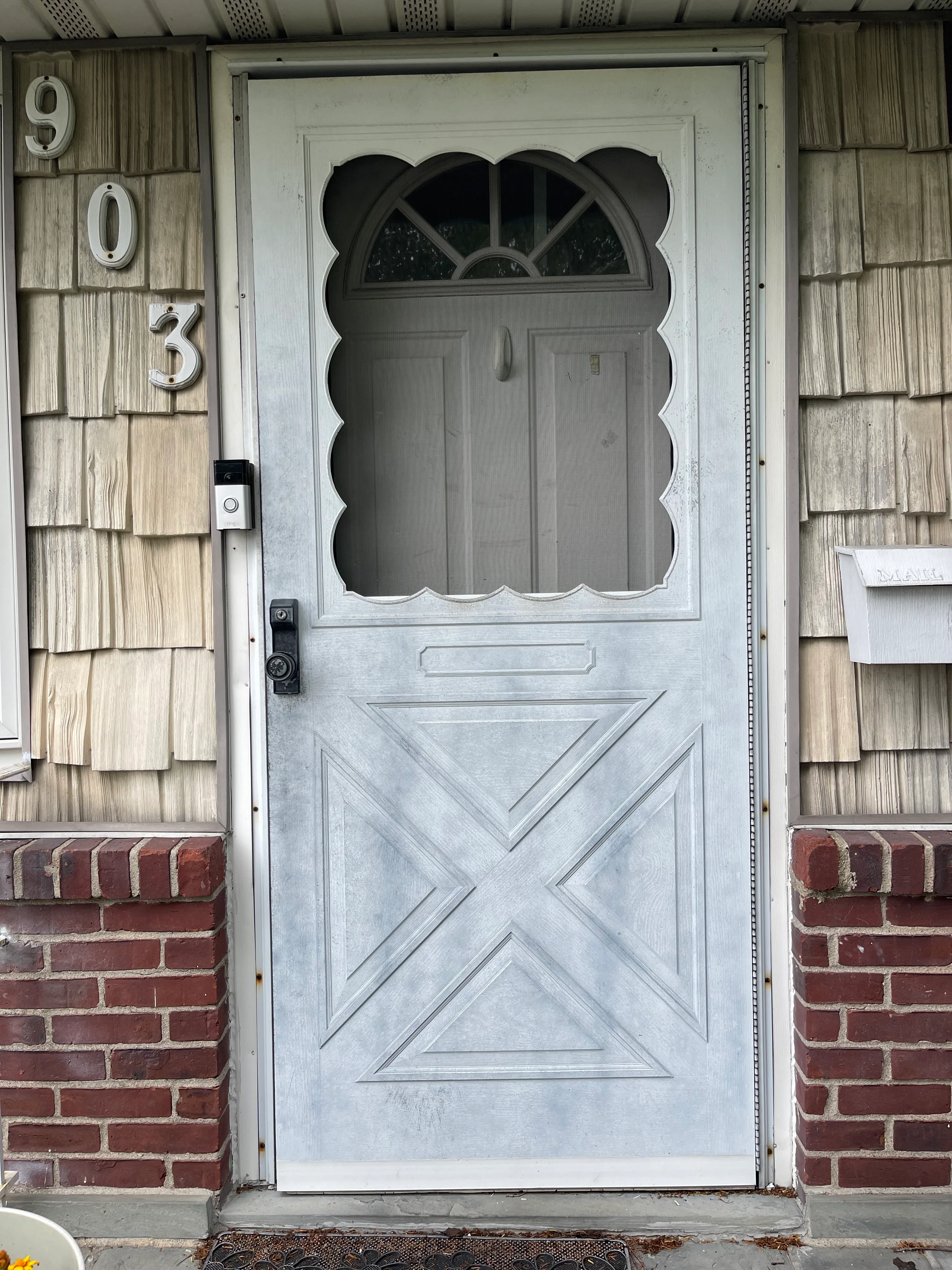White scalloped entry door with transom window and X-pattern panel on brick and cedar shake home